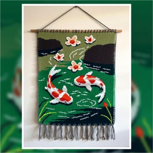 Può includere: Un arazzo da parete fatto a mano raffigurante una scena di stagno con due pesci koi. L'arazzo è principalmente verde, con pesci rossi e bianchi, fiori rosa e rocce nere. È rifinito con frange grigie e un'asta di legno per appenderlo.