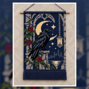 Op de afbeelding: Een rechthoekige wandhanger met een kruissteekontwerp van een zwarte raaf op een stenen richel. De afbeelding bevat een maansikkel, sterren, een hangende lantaarn en rode rozen tegen een donkerblauwe achtergrond met franjes aan de onderkant.