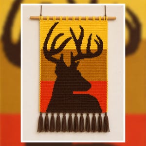 May include: A rectangular crochet wall hanging featuring a dark brown silhouette of a deer head with antlers against a horizontal striped background of yellow, tan, and orange. The piece hangs from a wooden dowel and has brown fringe tassels.