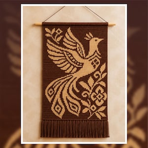 May include: A rectangular wall hanging featuring a dark brown and beige tapestry with a woven design of a bird with a long tail surrounded by floral elements. The piece is suspended from a wooden rod with a string and has a fringe at the bottom.