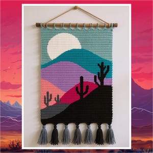 May include: A crocheted wall hanging featuring a desert scene with a white sun, mountains in teal, purple, pink, and black, and cactus silhouettes. It has grey and black tassels at the bottom and hangs from a wooden rod.