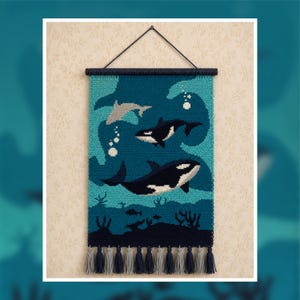 May include: A rectangular crochet wall hanging featuring two orcas and a dolphin swimming in deep blue and teal water. The design includes white bubbles and dark blue seaweed at the bottom. It hangs from a dark rod with alternating tassels.