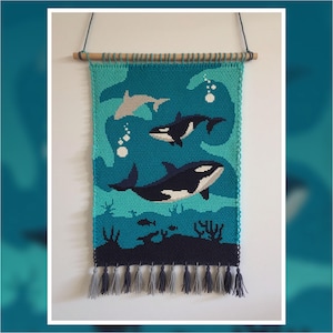 May include: A handcrafted wall hanging featuring an ocean scene with orcas, fish, and coral. The tapestry is primarily teal and blue, with a wooden rod and gray tassels at the bottom. The orcas are black and white.