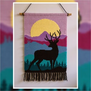 May include: A handcrafted wall hanging depicting a deer silhouette against a sunset scene. The tapestry uses shades of purple, yellow, teal, and black, and is finished with a fringe. It is suspended from a wooden dowel, creating a decorative piece.
