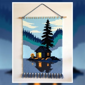 Könnte beinhalten: Ein gewebter Wandbehang mit einer Hüttenlandschaft. Das Kunstwerk zeigt eine Hütte mit einem beleuchteten Fenster, einen hohen Kiefernbaum und Berge, die sich im Wasser spiegeln. Die Farbpalette umfasst Blau-, Grün- und warme Gelbtöne von der Hütte.