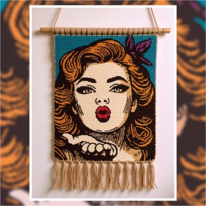 May include: A woven wall hanging featuring a pop art style image of a woman blowing a kiss. The woman has red hair, red lips, and a purple flower in her hair. The background is teal, and the piece is finished with tassels.