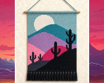 May include: A rectangular crochet wall hanging featuring a desert landscape with a large white sun, layered mountains in shades of blue, purple, and pink, and black silhouettes of saguaro cacti. The piece hangs from a wooden rod with black tassels.