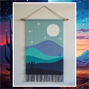 May include: A handmade wall hanging depicting a desert landscape. The tapestry shows a night sky with a full moon and stars above layered blue and teal mountains and cacti. The bottom edge has a fringe.