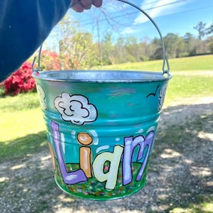 May include: A hand-painted teal metal bucket with the name "Liam" in colorful letters. The bucket features painted clouds, flowers, and a silver handle. The background shows a green lawn and trees.