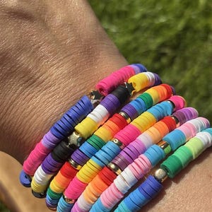 May include: A selection of vibrant beaded bracelets. The bracelets are made with flat, disc-shaped beads in a rainbow of colours, including pink, purple, blue, yellow, green, and orange. Gold-coloured spacers separate the beads. The bracelets are stacked on a wrist.