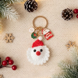 May include: A festive Christmas keychain featuring a crocheted Santa Claus with a red hat and white beard. The keychain includes gold-colored rings and charms, including a red tag that says "Xmas". The background includes pinecones, berries, and evergreen branches.