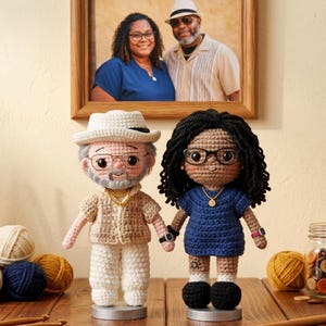 May include: Two handmade crochet dolls standing side by side. The left doll wears a beige shirt, white trousers, a hat, and glasses. The right doll has dark curly hair, wears a blue dress, glasses, and a necklace. Both dolls stand on circular bases.