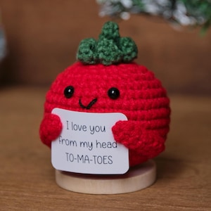 Op de afbeelding: Een handgemaakte rode gehaakte tomaat met een groene steel, met een bordje met de tekst "I love you from my head TO-MA-TOES". De tomaat heeft zwarte ogen en staat op een houten voet. Een kleine kerstboom is op de achtergrond te zien.