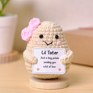 May include: A beige crocheted potato figure with a pink bow, black bead eyes, and a stitched smile. It holds a white rectangular sign that reads: Lil Tater, Just a tiny potato sending you a lot of love, with two small red hearts at the bottom.