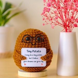 Puede incluir: Una figura de patata tejida a ganchillo de color marrón con gafas de plástico negro, sobre una pequeña base de madera. Sostiene un cartel rectangular blanco con el texto: Tiny Potato, I may be a tiny potato, but I believe your dreams can grow big.
