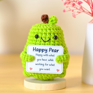 May include: A lime green crocheted pear figurine with a brown stem, black bead eyes, and a stitched smile. It holds a white rectangular sign that reads: Happy Pear, Happy with what you have, while working for what you want, with two small pink hearts.