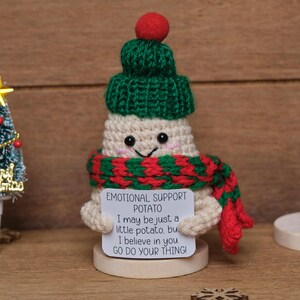 Op de afbeelding: Een gehaakte aardappel figuur met een groene muts, rood-groene sjaal en een bord met de tekst "EMOTIONAL SUPPORT POTATO". Een kleine kerstboom en houten sneeuwvlokken staan ook in de afbeelding.