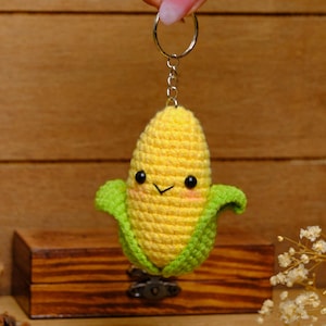 Könnte beinhalten: Ein handgefertigter Maiskolben-Schlüsselanhänger mit einem lächelnden Gesicht. Der Mais ist gelb mit grünen Blättern und schwarzen Augen. Der Schlüsselanhänger ist an einem silbernen Metallring befestigt.
