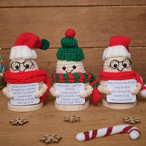 May include: Three handmade crochet potato figures wearing hats, scarves, and glasses. Each potato holds a sign with a holiday message. A small Christmas tree, snowflakes, and a candy cane are also visible.