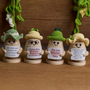 Op de afbeelding: Vier handgemaakte gehaakte aardappel figuren, elk met een unieke hoed en een klein bordje. De bordjes tonen verschillende berichten, waaronder "You look AMAZING Kelly!" en andere inspirerende citaten. De aardappelen staan op houten voetstukken.