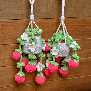 May include: Two handmade crochet hanging planters. Each planter is a beige pot with green leaves and white flowers. Bright pink crochet strawberries hang below. The planters are suspended by white cords against a wooden background.