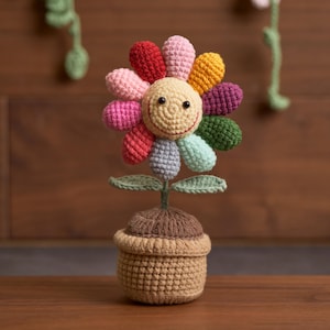 May include: A colorful crocheted flower in a pot. The flower has a smiling face and petals in various colors, including red, pink, yellow, and green. The pot is a light brown color. The flower is a handmade item.