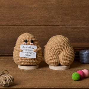 Puede incluir: Dos figuras de patata de ganchillo, una con un cartel que dice "Life was all out of lemons, so here's a potato instead." Las patatas son de color canela con mejillas rosas y ojos negros. Están sobre bases de madera.