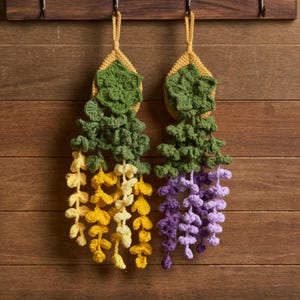 May include: Two handmade crochet wisteria flower wall hangings. One features yellow and cream-colored flowers, the other purple. Both have green leaves and a yellow top with a loop for hanging. The background is a wooden wall.