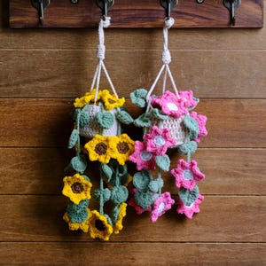 May include: Two handmade crochet hanging planters. One features yellow sunflowers with green leaves, the other pink flowers with green leaves. Both are suspended by white cords from a wooden rack, showcasing a rustic, handcrafted aesthetic.