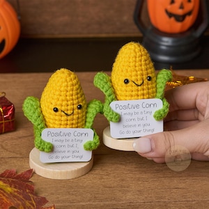 Könnte beinhalten: Zwei handgefertigte gehäkelte Maisfiguren mit lächelnden Gesichtern und grünen Armen, die jeweils ein Schild mit der Aufschrift "Positive Corn" halten. Die Schilder enthalten den Text "I may be a tiny corn, but I believe in you. Go do your thing!"