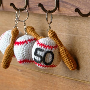 May include: Crocheted baseball keychains hanging on a wooden surface. Each keychain features a white baseball with red stitching and a small brown bat. One baseball displays the number 50 in black.