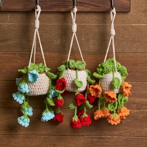May include: Three handmade crochet hanging planters with cream-colored pots and trailing flowers in blue, red, and orange. The planters are suspended by cream-colored cords from hooks on a wooden background. The crochet work is detailed and colorful.