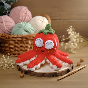 Op de afbeelding: Een handgemaakt, gehaakt octopus speelgoed in rood en wit met een aardbei-vormige kop en groene steel. De octopus staat op een houten schijf, met een houten haaknaald en garen op de achtergrond. Het speelgoed heeft witte ogen en een lachend gezicht.