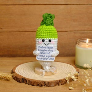 May include: A crocheted radish figurine with a green top, white body, and a smiling face. The radish holds a sign that reads "Positive Radish I may be a tiny Radish but I believe in you Go do your thing!" It sits on a wooden base.