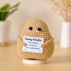Op de afbeelding: Een beige gehaakt aardappelfiguurtje met zwarte plastic ogen en roze geborduurde wangen. Het staat op een kleine ronde houten voet en houdt een wit rechthoekig bordje vast met de tekst: Funny Potato, Life was all out of lemons, so here's a potato intead.