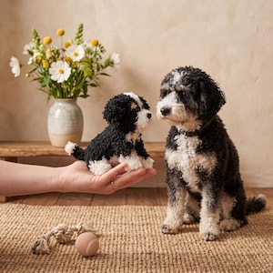 May include: A small, handcrafted plush dog figurine made of black and white yarn, featuring a textured, curly appearance. The miniature toy is held in a palm, positioned near a live black and white dog with similar curly fur and a dark collar.