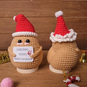 Op de afbeelding: Twee gehaakte kerstversieringen in de vorm van aardappelen. Elke aardappel draagt een rode kerstmuts met een witte rand. Eén aardappel houdt een bordje vast met de tekst "Christmas Potato Merry Christmas". De aardappelen zijn bruin en staan op houten voetstukken.