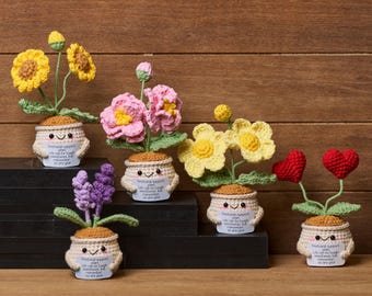 May include: A collection of handmade crochet flower arrangements in small pots. The flowers are yellow, pink, purple, and red. Each pot has a smiling face and a small tag with text. The background is a wooden surface.