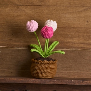 Puede incluir: Arreglo floral de ganchillo con tres tulipanes en una maceta marrón. Los tulipanes son rosas, blancos y fucsias, con tallos y hojas verdes. La maceta es marrón con un borde festoneado, sobre un fondo de madera.