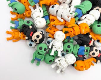May include: A collection of small, 3D-printed plastic figurines featuring various characters including skeletons, aliens, and figures with black hair, rendered in solid colors of white, orange, green, blue, and black, scattered across a white surface.