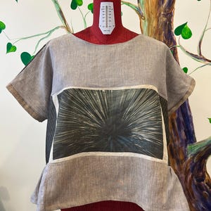 Puede incluir: Una blusa de lino gris con cuello redondo y mangas cortas. La blusa presenta un panel rectangular con un diseño gráfico en verde oscuro y blanco. La prenda se exhibe en un maniquí rojo.