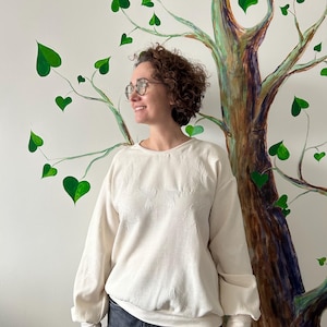 May include: Cream-colored sweatshirt with a subtle floral pattern. The long-sleeved top has a crew neck and a relaxed fit. The person in the image is wearing dark wash jeans. The background features a painted tree with green heart-shaped leaves.