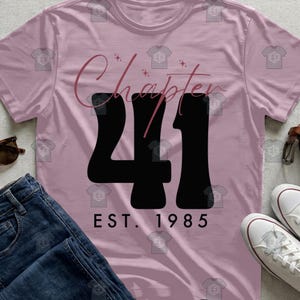Puede incluir: Una camiseta rosa claro con las palabras "Chapter 41" en fuente cursiva y grandes números negros. Debajo de los números, el texto "EST. 1985" está impreso en una fuente sencilla. La camiseta se muestra con vaqueros azules y zapatillas blancas.