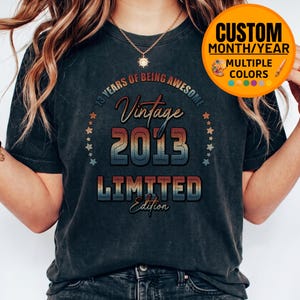 Peut inclure: Un t-shirt gris foncé à manches courtes avec un motif imprimé comprenant le texte 13 YEARS OF BEING AWESOME, Vintage, 2013 et LIMITED Edition dans une police rétro aux tons dégradés rouge, orange et bleu, entouré de petites étoiles.