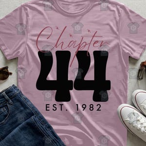 Puede incluir: Una camiseta rosa claro con las palabras "Chapter 44" en fuente cursiva y grandes números negros. Debajo de los números, el texto "EST. 1982" está impreso. La camiseta se combina con vaqueros azules y zapatillas blancas.