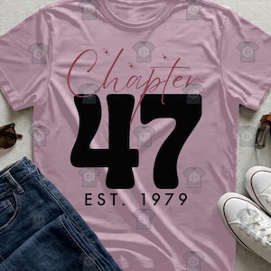 May include: A light pink t-shirt with the words "Chapter 47" in a cursive font and the number "47" in bold black. Below the number is "EST. 1979" in a simple font. The shirt is paired with blue jeans and white sneakers.