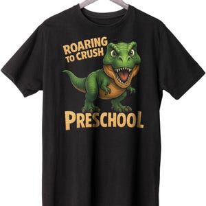 May include: Black t-shirt featuring a green and orange cartoon dinosaur. The text "ROARING TO CRUSH" is above the dinosaur, and "PRESCHOOL" is below. The shirt is displayed on a wooden hanger.