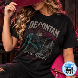 Könnte beinhalten: Ein schwarzes T-Shirt mit einer Grafik eines Skeletts, das ein Bandana und eine Schürze trägt und an einem Waschbecken arbeitet. Der Text lautet DECONTAM AFTER DARK, DIRTY IN CLEAN OUT und Nothing Scares Me Anymore. Das Design zeigt kleine Fledermäuse.