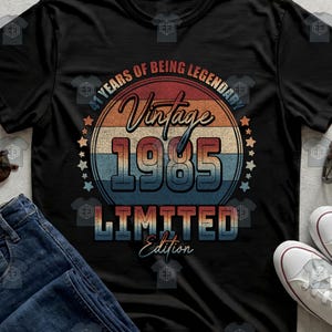 May include: A black t-shirt featuring a circular graphic with horizontal stripes in red, beige, and blue. The text reads 41 YEARS OF BEING LEGENDARY, Vintage, 1985, and LIMITED Edition. Small stars are arranged along the sides of the circle.