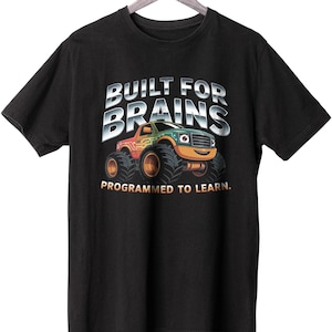 May include: Black t-shirt with the text "BUILT FOR BRAINS" above a colorful monster truck graphic. The truck is green, orange, and yellow with large black tires. The text "PROGRAMMED TO LEARN" is below the truck.